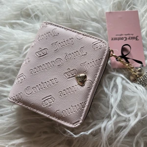Ljusrosa plånbok från Juicy Couture - Supersöt ljusrosa plånbok från Juicy Couture med präglad logga och små kronor över hela utsidan. Plånboken har guldfärgade detaljer, dragkedja och flera kortfack. Insidan är fodrad med mönstrat tyg och har plats för både kort och mynt.