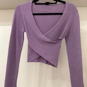 Ljuslila omlottopp - Superfin ljuslila omlottopp med croppad passform och långa ärmar. Toppen är ribbstickad och har en djup v-ringning som ger en snygg och trendig look. Perfekt att styla med höga jeans eller kjol! Dessvärre har toppen 2 defekter (se två sista bilderna) som dock knappt syns när man har den på sig.