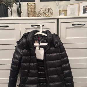 Svart Moncler pufferjacka med huva (bady)  - Säljer en svart pufferjacka från Moncler med glansig finish och huva. Jackan har dragkedja framtill, två fickor med dragkedja och klassisk Moncler-logga på insidan. Perfekt för kalla dagar och riktigt snygg streetstil. Använd några gånger, som ny. Äkta såklart! 