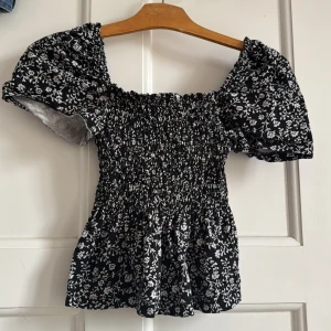 Svart blommig topp från H&M XS - Supersöt svart topp från H&M i storlek XS med vitt blommönster. Modellen har puffiga korta ärmar, smockad överdel och lätt utställd nertill. Perfekt för dig som gillar romantisk stil och vill ha något som sticker ut lite extra.