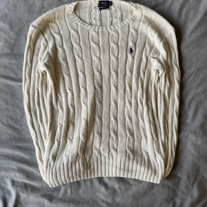 Vit kabelstickad tröja Polo Ralph Lauren - Kabelstickad vit tröja från Polo Ralph Lauren i mjuk pima bomull. Klassisk rund halsringning, ribbade muddar och ikonisk broderad logga på bröstet. Perfekt för lager-på-lager och snygg till jeans eller kjol.