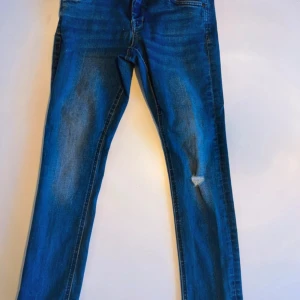 Blå jeans från Name It, stl 146 - Snygga blå jeans från Name It i storlek 146. Klassisk femficksmodell med dragkedja och knapp framtill. Jeansen har raka ben och lätt slitna detaljer på framsidan för en cool look. Perfekta till vardagsstilen!