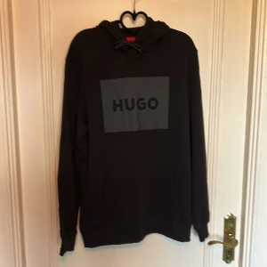 Hugo Boss - Hoodie - Svart hoodie från Hugo Boss med stor logga framtill. Klassisk huva med snörning och känguruficka. Mjuk bomullskänsla och långa ärmar, perfekt för en chill och stilren look.