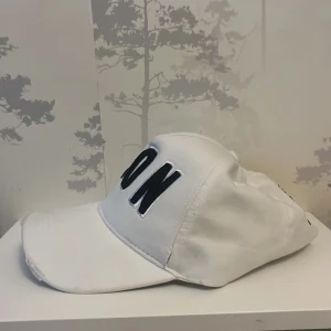 Vit keps från Dsquared2 med brodyr - Snygg vit keps från Dsquared2 med stor svart brodyrtext 'ICON' framtill och Dsquared2 broderat på sidan. Justerbar rem baktill för perfekt passform. Klassisk böjd skärm och cool streetstil. Perfekt accessoar för att lyfta din outfit.Priset ej hugget !