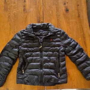 Svart dunjacka från Polo Ralph Lauren - Säljer en svart dunjacka från Polo Ralph Lauren med klassisk quiltad design och dragkedja framtill. Jackan har två fickor med dragkedja och en liten röd logga på bröstet. Perfekt för kyliga dagar och riktigt snygg till jeans eller hoodie. Passar till någon som är 160-165cm lång. Är i storlek L (Barnstorlek) men passar som en xs.