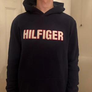Mörkblå hoodie från Tommy Hilfiger - Säljer en mörkblå hoodie från Tommy Hilfiger med stor broderad logga i vitt och rött framtill. Tröjan har huva, lång ärm och en klassisk känguruficka. Perfekt för dig som gillar streetwear och vill ha en stilren look.