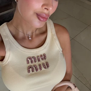 Beige ribbad linnetopp från Miu Miu - Supersnygg beige ribbad linnetopp från Miu Miu med glittrande rosa och silvriga pärldetaljer på bröstet. Toppen är ärmlös och har en croppad passform som ger en fräsch och trendig look. Perfekt att styla med jeans eller kjol för en cool vibe.