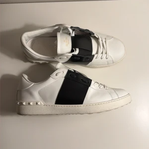 Valentino Garavani Open sneakers - Säljer ett par vita Valentino Garavani Open sneakers i skinn med bred svart rem över mitten och ikoniska nitar på hälen. Klassisk rund tå, platt sula och vita snören. Perfekta för dig som vill ha en clean men ändå lyxig look. Skick 9/10 storlek 45