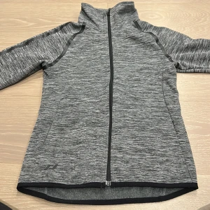 Grå träningsjacka från Pro Touch - Sportig gråmelerad träningsjacka från Pro Touch med hel dragkedja, hög krage och tumhål i ärmsluten. Jackan är tillverkad i mjuk polyester och har svarta detaljer längs ärmarna. Perfekt för aktiva dagar och träning.