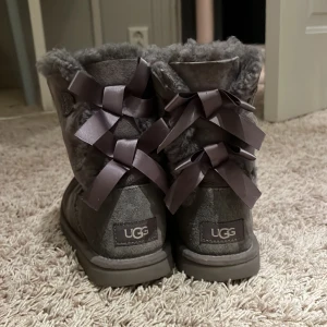 Grå UGG boots med rosetter - Mysiga grå UGG boots i mocka med fluffigt foder och dekorativa satinrosetter baktill. Skorna har platt sula och rund tå, perfekt för kalla dagar. Klassisk UGG-logga bak och grovt mönstrad yttersula för bra grepp.