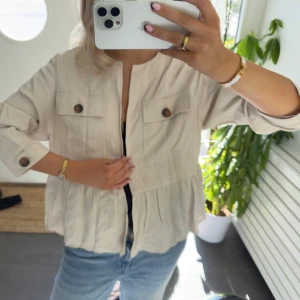Beige overshirt med knappar - Snygg beige overshirt med volang nedtill och stora bruna knappar på fickorna och ärmsluten. Jackan har en loose passform, långärmade ärmar och två bröstfickor. Perfekt att slänga över en topp för en chill look.