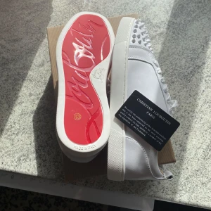 Christian Louboutin vita sneakers med nitar - Säljer ett par vita sneakers från Christian Louboutin med ikonisk röd sula och coola nitar på tån. Skorna har snörning och är tillverkade i skinn med en platt sula. Perfekta för dig som vill sticka ut med exklusiv design och edgy detaljer.