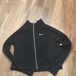 Svart zip-tröja från Nike - Svart långärmad zip-tröja från Nike med vit logga på bröstet. Tröjan har ribbad struktur, hög krage och hel dragkedja framtill. Materialet är en blandning av bomull, polyester och elastan.