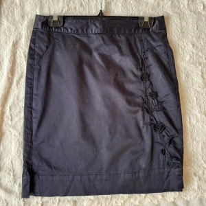 Svart pennkjol med broderi från Jackpot - Snygg svart pennkjol från Jackpot med broderade blommor längs ena sidan. Kjolen har en klassisk, figurnära passform och är tillverkad i bomullsmix med dragkedja bak. Lite glansigt material, underkjol i polyesterliknade tyg. Perfekt för dig som gillar stilrena detaljer och vill ha en kjol som sticker ut lite extra. Pennkjol är tillbaka hösten 25  som trend enligt Elle. :)