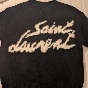 Svart stickad Saint Laurent tröja - Cool svart stickad tröja från Saint Laurent med stor vit, fluffig logotyp framtill. Tröjan har rund halsringning och ribbade muddar. Perfekt för dig som vill sticka ut med en statement piece.