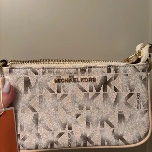 Vit axelväska Michael Kors logomönster - Snygg vit axelväska från Michael Kors med ikoniskt MK-logomönster över hela väskan. Väskan har guldfärgade detaljer, dragkedja upptill och justerbar axelrem. Tillverkad i tåligt syntetmaterial och perfekt för dig som vill ha en trendig och stilren accessoar. Väskan är helt oanvänd men inte äkta