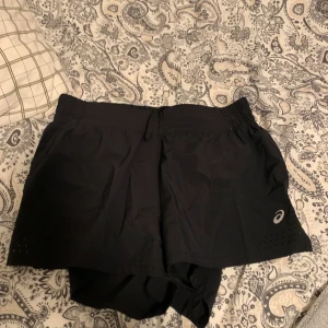 Svarta träningsshorts från Asics XS - Svarta träningsshorts från Asics i storlek XS. De har elastisk midja med snörning, reflekterande detaljer och innerbyxa. Perfekta för löpning eller gym, med lätt och snabbtorkande material. Klassisk sportig look med Asics-logga på benet och en mindre bakficka. Köpte i USA. Köpta för runt 400kr använd 1 gång. Mitt pris 200kr