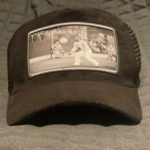 Svart keps från Lozama Platinum Edition - Snygg svart keps från Lozama Platinum Edition med mesh-bakstycke och justerbar rem. Framtill finns ett svartvitt tryck med tennismotiv som ger en unik vibe. Kepsen levereras med originalpåse från Lozama Headwear. Kostar 550 Ny använd ungefär 7 gånger väldigt fräsch