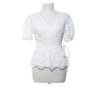 Vit spetsblus med knyt från Lipsy - Superfin vit blus från Lipsy med v-ringning, kort puffärm och omlottknytning i midjan. Blusen är täckt av blommig spets och har en lätt volangkant nedtill. Perfekt för dig som gillar romantiska detaljer och vill ha en feminin look.