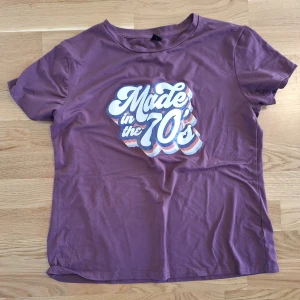Lila t-shirt med 70-tals tryck - Retroinspirerad lila t-shirt med coolt 'Made in the 70's'-tryck i vitt, blått och rosa på bröstet. Klassisk rund hals och korta ärmar. Perfekt för dig som gillar vintagevibbar och mjuk bomullskänsla.
