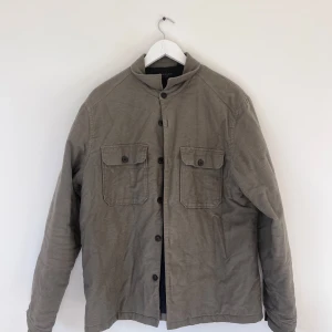 Olivgrön/grå overshirt från AllSaints - Olivgröngrå overshirt från AllSaints i storlek L. Jackan har två bröstfickor med lock och knappar, klassisk krage och knäppning framtill. Tillverkad i mjuk bomull med lätt struktur, perfekt för lager-på-lager. Snygg och enkel att styla till det mesta.