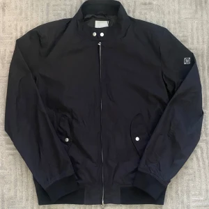 Svart jacka från J.Lindeberg - Stilren svart jacka från J.Lindeberg med klassisk passform. Jackan har dragkedja framtill, ribbade muddar och två fickor med tryckknappar. Den klassiska metallbadgen med logga på ärmen ger en exklusiv touch. Storlek L men den passar både L och M, vid fler frågor eller funderingar tveka inte på att skriva!