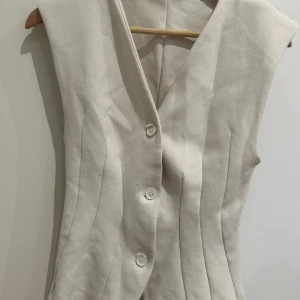 Beige väst från Zara med knappar - Snygg beige väst från Zara i storlek M. Västen har en figurnära passform med markerade sömmar och tre knappar framtill. Perfekt att styla över en skjorta eller t-shirt för en trendig look.