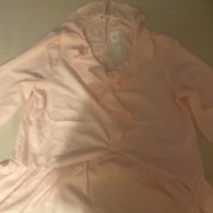 Ljusrosa omlotttröja  med volang - Supersöt ljusrosa klänning med omlott och volangdetaljer. Klänningen har långa ärmar och en mjuk, flowy känsla. Perfekt för dig som gillar romantiska och feminina plagg. Passar till vår och sommar!