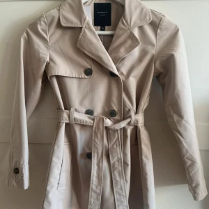 Beige trenchcoat från Name It - Stilren beige trenchcoat från Name It med klassisk dubbelknäppning, bälte i midjan och stora knappar. Jackan har långa ärmar, två fickor framtill och en snygg krage. Perfekt för dig som vill ha en tidlös och trendig look.