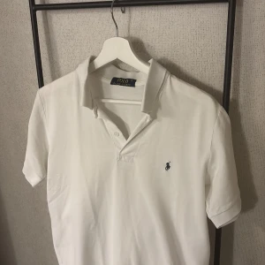 Vit polo t-shirt från Ralph Lauren - Vit klassisk polo t-shirt från Ralph Lauren med korta ärmar och krage. Ikoniska broderade loggan på bröstet. Tillverkad i mjuk bomull som känns skön mot huden. Perfekt för dig som gillar stilrena och tidlösa plagg. Storlek M. 