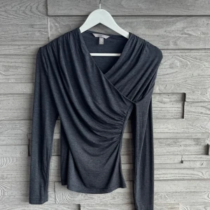 Mörkgrå omlottopp från H&M - Snygg mörkgrå omlottopp från H&M i stretchigt material. Toppen har långa ärmar, draperad effekt framtill och en smickrande v-ringning. Använd en gång.