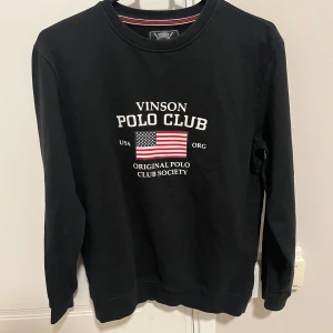 Svart sweatshirt Vinson Polo Club - Svart sweatshirt från Vinson Polo Club med tryck av amerikansk flagga och text på bröstet. Tröjan har rund halsringning, långa ärmar och är tillverkad i mjuk bomull. Perfekt för en sportig och avslappnad stil.
