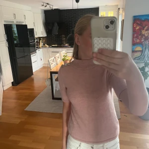 Rosa stickad tröja med knappar - Supersöt ljusrosa stickad tröja med korta ärmar och ribbad hög krage. Snygga silvriga knappar på ena axeln ger en unik touch. Perfekt till jeans eller kjol för en chill men ändå uppklädd vibe.