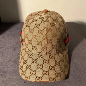 Keps med monogram och ränder - Säljer en keps med klassiskt GG-monogram över hela kepsen och gröna/röda ränder på sidorna. Kepsen är tillverkad i en blandning av bomull och merinoull, med justerbar rem baktill och detaljer i brunt. Perfekt för dig som vill sticka ut.