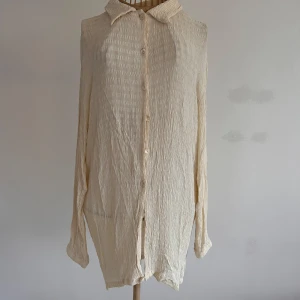 Krämvit skjorta från Vero Moda, stl L - Krämvit skjorta från Vero Moda i storlek L. Skjortan har en snygg krinklad struktur, klassisk krage och knäppning framtill. Tillverkad i 100% viskos för en lätt och luftig känsla. Perfekt för dig som gillar en stilren och avslappnad look.