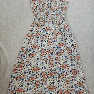 Blommig långklänning från SHEIN - Supersöt långklänning från SHEIN med blommigt mönster i rött, blått och grönt på vit botten. Klänningen har smockad överdel, volangärmar och rund hals. Materialet är mjukt och lätt, perfekt för varma dagar. Passar dig som gillar romantisk och feminin stil.