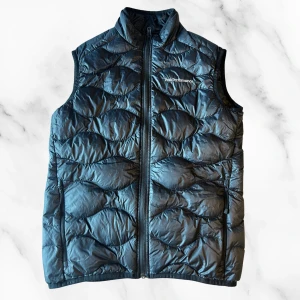 Svart dunväst från Peak Performance - Svart dunväst från Peak Performance, modell Helium Down Vest. Västen har quiltad design med vågigt mönster, hög krage och dragkedja framtill. Perfekt för lager-på-lager och snygg till både hoodie och t-shirt.