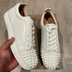 Vita Christian Louboutin sneakers(prisförslag) - Säljer ett par vita sneakers från Christian Louboutin i skinn med ikoniska röda sulor och coola vita nitar framtill. Skorna har snörning och en clean, modern look som sticker ut. Perfekta för dig som vill ha något extra och vågar sticka ut i mängden.