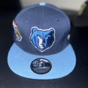 Memphis Grizzlies snapback keps New Era - Snygg snapback keps från New Era med Memphis Grizzlies-logga framtill och NBA-logga bak. Kepsen är mörkblå med ljusblå skärm och justerbar baksida. Broderade detaljer och klassisk 9FIFTY-modell. Perfekt för basketfans!