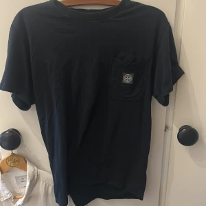 Svart Stone Island t-shirt med ficka - Svart t-shirt från Stone Island med klassisk logotyp på bröstfickan. T-shirten har rund hals, korta ärmar och är tillverkad i mjuk bomull. Perfekt för dig som gillar streetwear och vill ha en stilren look.