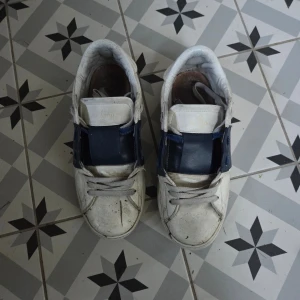 Valentino Garavani vita och blå sneakers - Säljer ett par snygga Valentino Garavani sneakers i vitt och mörkblått skinn. Skorna har en bred blå skinnpanel över mitten, klassisk snörning och rund tå. Perfekta för dig som gillar exklusiva och stilrena sneakers med en lyxig känsla.