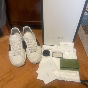 Gucci Ace vita sneakers med gröna detaljer - Gucci Ace sneakers i vitt skinn med klassiska gröna och röda ränder på sidorna och grön krokopräglad häl med Gucci-logga. Platt sula, rund tå och snörning framtill. Kommer med originalkartong, extra skosnören och dustbag samt kvitto. Byte mot Valentino open är av intresse, gärna i storlek 40,5/41 och i färgen navy men svart funkar också, bara hör av er!