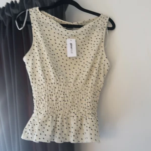 Prickig ärmlös topp från Gina Tricot - Supersöt ärmlös topp från Gina Tricot i beige med svarta prickar. Toppen har smockad midja som ger en snygg form och lätt volang nertill. Perfekt för varma dagar och enkel att styla med jeans eller kjol.