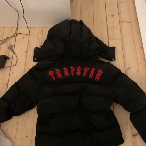 Svart pufferjacka från Trapstar - Säljer en svart pufferjacka från Trapstar med stor röd logga på ryggen den missar en knapp på luvan som syns på bilden den är äkta å har kvitto som kan visas vid intresse priset kan förhandlas vid snabb affär bara å fråga om fler bilder om de skulle velas 