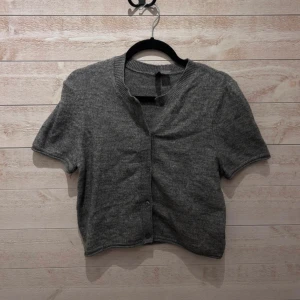 Grå kort kofta från H&M Divided - Kort grå kofta från H&M Divided med rund hals och knappar framtill. Koftan har ribbade detaljer vid hals och knappslå samt korta ärmar. Perfekt att slänga över en topp för en chill look. Mjuk stickad känsla.