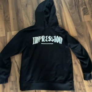 Svart hoodie med tryck 'IMPRESSION' - Svart hoodie med stor huva och vitt tryck på bröstet där det står 'IMPRESSION' i gotisk stil. Under texten finns även mindre text. Hoodien har långa ärmar och breda muddar vid ärmslut och nederkant. Perfekt för en chill och avslappnad look. Kom med bud