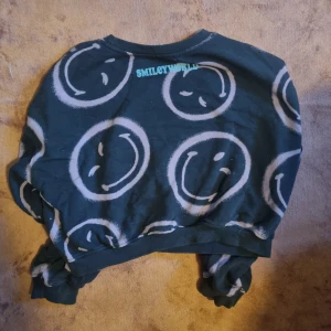 Svart croppad tröja SmileyWorld - Cool svart croppad tröja från SmileyWorld med stora vita smiley-mönster över hela. Tröjan har rund halsringning och långa ärmar med muddar. Perfekt för dig som gillar streetstyle och vill sticka ut lite extra.