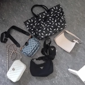 Paket med 5 väskor - Säljer fem snygga väskor: en stor svart shoppingväska med fjädermönster, en ljusbeige axelväska i skinn, en vit quiltad handväska med kedja, en blå axelväska med monogram och en svart axelväska i nylon med dragkedja och extra liten pouch. Pr*adans dragkedja funkar ej, annars ska de vara hela, men i behov av en tvätt. Säljer billigt pga skicket och jag rensar hemma.