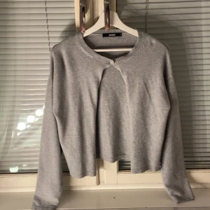 Grå croppad kofta från Bik Bok - Grå croppad kofta från Bik Bok med rund halsringning och en knapp framtill. Långärmad och i mjukt material som passar perfekt till lager-på-lager. Enkel och stilren design som funkar till det mesta.