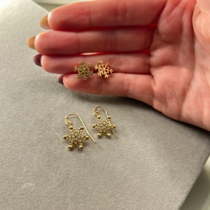 Guldiga snöflingeörhängen i två modeller - Två par örhängen i form av snöflingor i guldig färg. Ena paret är små studs och det andra paret är hängande krokar. Båda har detaljerad snöflingeform och ger en vintrig vibe. Perfekta för dig som vill sticka ut med ett unikt och lekfullt smycke.